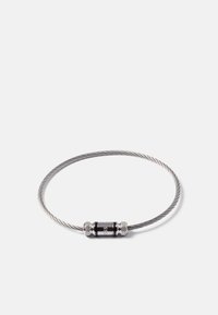 Bracelet en câble métallique argenté fin et torsadé, avec un fermoir cylindrique noir gravé d'un petit logo sur fond blanc.