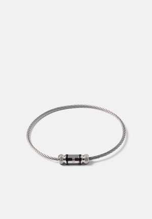 Bracelet en câble métallique argenté fin et torsadé, avec un fermoir cylindrique noir gravé d'un petit logo sur fond blanc.