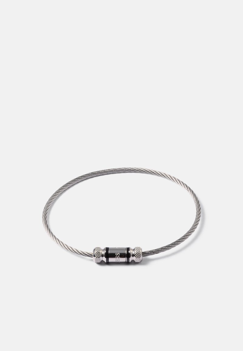 Bracelet en câble métallique argenté fin et torsadé, avec un fermoir cylindrique noir gravé d'un petit logo sur fond blanc.