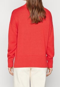 Rode gebreide trui met een hoge hals, geribbelde manchetten en een geribbelde zoom. Het heeft een gestructureerde textuur en een losse pasvorm. Draagt over lichtgekleurde broek.