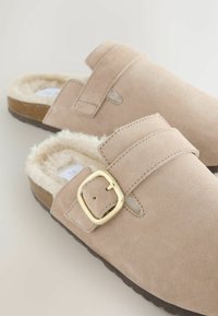 Zuecos de ante beige claro, tipo slip-on, con forro de pelo suave, correa ajustable con hebilla y suela marrón texturizada.