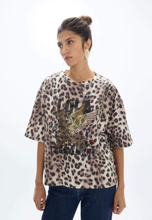 Mujer con el pelo recogido lleva una camisa de gran tamaño con estampado de leopardo y gráfico de un animal alado y vaqueros oscuros, de pie frente a un fondo blanco.