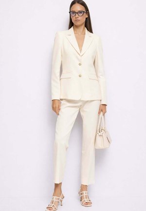 Donna che indossa un blazer crema su misura e pantaloni abbinati, tiene una borsa crema, indossa occhiali e sandali con tacco aperti.