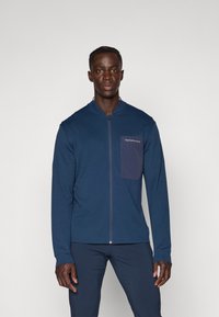 Marineblaue Zip-Jacke mit langen Ärmeln, ausgestattet mit einer Brusttasche in einem dunkleren Farbton. Glattes Material mit einem sanften Glanz und minimaler Markenkennzeichnung.