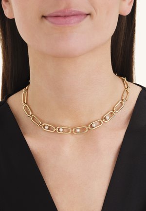 Collier chaîne en or avec de grands maillons rectangulaires, chacun contenant une petite perle blanche, porté avec un haut noir.