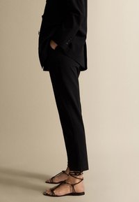 Mustat räätälöidyt housut, joissa on suora leikkaus, yhdistettyinä mustiin nauhapestäviin sandaaleihin, joissa on litteä pohja ja ohuet remmit, esiteltyinä neutraalilla taustalla.