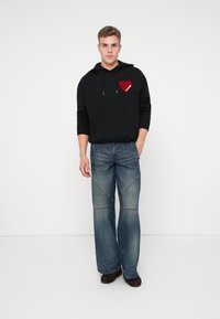 Czarna bluza z kapturem z graficznym motywem czerwonego serca i tekstem, zestawiona z jasnoniebieskimi dżinsami w stylu dzwonów z detalami szwów. Modelka stoi na jednolitym tle.