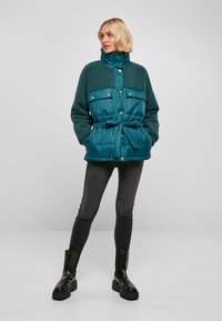 Veste matelassée turquoise avec un haut en polaire vert doux, taille ceinturée, boutons-pression et col haut ; associée à un jean noir ajusté et des bottes épaisses.