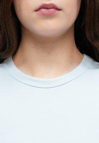 Svjetloplava, rebrasta majica s okruglim izrezom. Tkanina je glatka. Istaknuti detalji uključuju šavove oko izreza i bez vidljivih uzoraka.