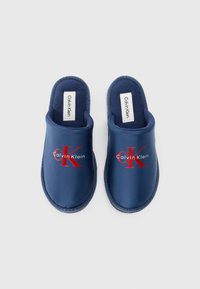 Námořnické modré pantofle s hladkým, matným povrchem. Obsahují červené logo "CK" a text "Calvin Klein". Otevřené zadní provedení a vycpaná stélka pro pohodlí.
