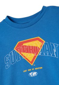 Blauwe T-shirt met een rood en geel Superman schild logo, witte omlijnde "SUPERMAN" tekst, en gele "LAATSTE ZOON VAN KRYPTON" tekst boven een wereldbol icoon.