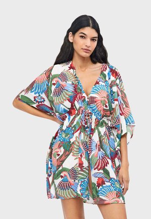 Mujer de cabello largo y oscuro, vestida con un vestido corto y suelto con estampados vibrantes de aves tropicales y flores, de pie con una mano en la cadera.