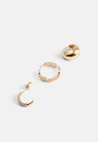 Tři zlaté náušnice: lesklý tvar kopule, texturovaný kroužek s malými kamínky a zakřivený design s třpytivými akcenty.