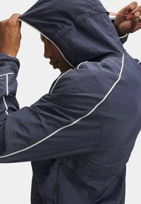 Veste légère navy avec capuche, dotée d'un tissu texturé, d'accents réfléchissants blancs le long des coutures et d'un cordon de serrage ajustable à la capuche.