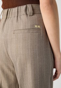 Beige randiga byxor med en slät textur, med en bakficka och guldbroderade initialer "M.M." vid midjan.