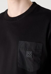 Gros plan d'une chemise noire avec une poche poitrine texturée présentant le nom de la marque en relief "HUGO" en noir sur noir.