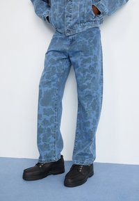 Blå denimbukser med et mønstret design, der har en afslappet pasform. Sammen med sorte sko og en matchende jakke med knapdetaljer.