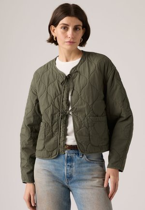 Frau trägt olivgrüne gesteppte Jacke, vorne gebunden, weißes Hemd, hellblaue Jeans und goldene Creolen, steht vor einfarbigem Hintergrund.