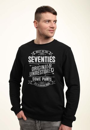 Mand med kort hår iført sort sweatshirt med teksten "Bygget i halvfjerdserne, Original og urestaureret, Nogle dele stadig i funktion," stående.