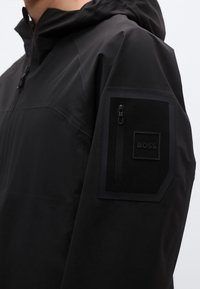 Fekete vízálló dzseki cipzáras zsebbel az ujján, texturált anyagból, és egy feltűnő márkajelzés fekete folttal.