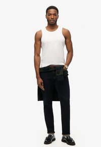 Camiseta de algodón blanca sin mangas, jeans de mezclilla oscuros y zapatos negros. Un suéter verde atado a la cintura añade un detalle adicional.