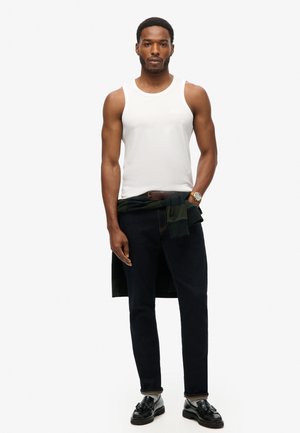 Un débardeur en coton blanc, un jean en denim foncé et des chaussures noires. Un pull vert noué autour de la taille ajoute un détail supplémentaire.