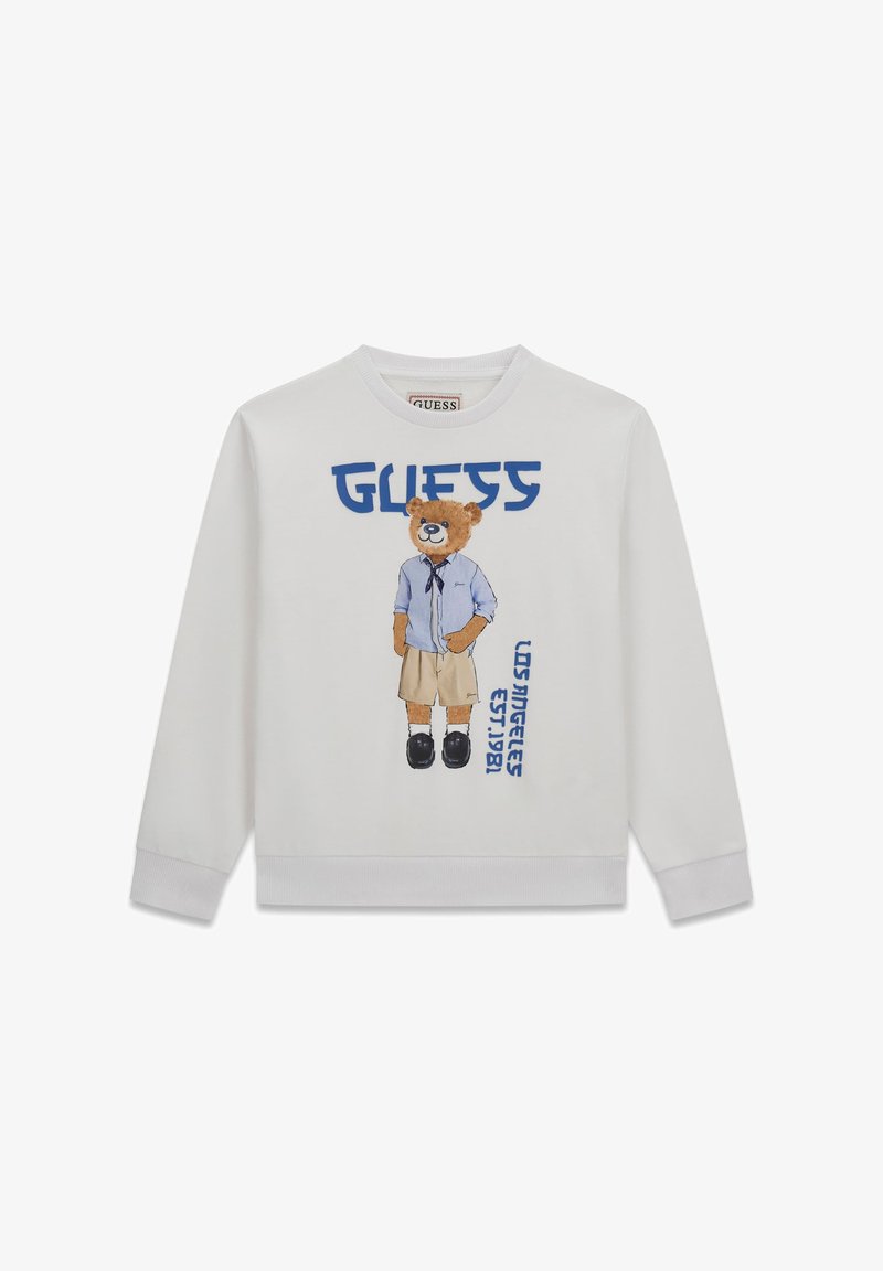 Witte sweatshirt met een teddybeer die een blauw shirt, beige korte broek, zwarte schoenen en blauwe tekst draagt met de woorden "GUESS" en "Los Angeles Est. 1981".
