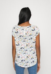 Blusa floreale in tessuto leggero con una base bianca e motivi multicolori in blu, giallo e rosa. Scollo rotondo e dettaglio con zip sul retro.