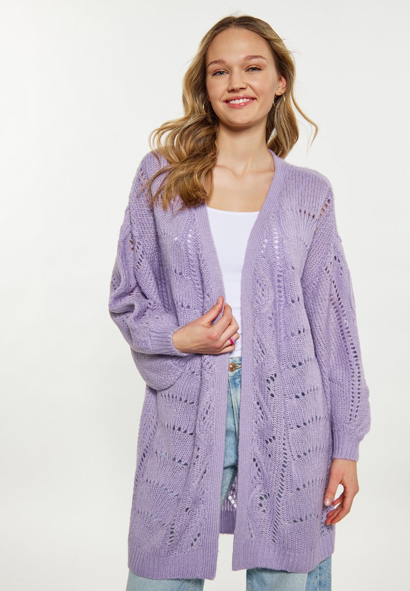 Lila stickad cardigan med öppen framsida, vida ärmar och detaljerat mönster i öppet arbete; matchas med en vit topp och ljusblå jeans.