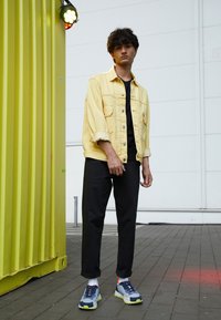 Giacca di jeans gialla sopra una maglietta nera, pantaloni neri arrotolati e sneakers multicolori con suola testurizzata, in piedi contro un muro giallo.