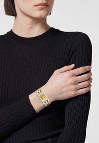 Pulsera de oro y plata con una cara rectangular que presenta un emblema de león, colocada sobre un suéter negro con canalé de una modelo.