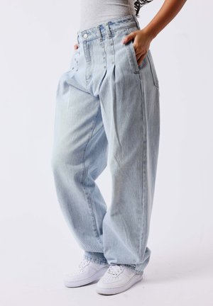 Personne portant un jean en denim bleu clair ample et des baskets blanches, debout les mains dans les poches devant un fond uni.