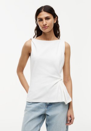 Femme portant un haut blanc sans manches avec un nœud sur le côté et un jean bleu clair, debout avec une main dans la poche devant un fond uni.