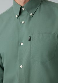 Camicia verde con bottoni, realizzata in tessuto liscio, con colletto a punta, bottoni bianchi e una tasca sul petto sinistro con una piccola etichetta logo.
