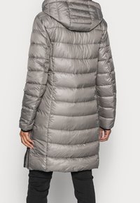 Długa srebrnoszara pikowana kurtka puchowa z kapturem, noszona przez osobę widoczną z tyłu, zestawiona z czarnymi spodniami.