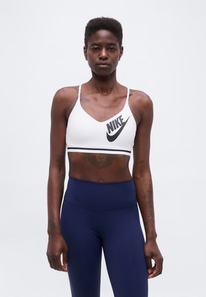 Persoană atletică cu păr scurt, purtând un sutien sport alb Nike și colanți bleumarin, stând pe un fundal alb simplu.