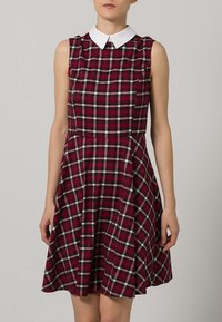 Červené kostkované šaty bez rukávů s bílým límečkem, přiléhavým vrškem a rozšířenou sukní. Vyrobeno z měkké tkaniny s klasickým tartanovým vzorem.
