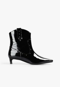 Botín de piel de charol negra con punta puntiaguda, tachuelas decorativas y tacón bajo. Presenta un diseño estructurado y una lengüeta para facilitar su colocación.