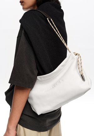 Osoba w czarnej koszuli i beżowych spodniach niesie dużą białą torbę na ramię z wzorzystym paskiem ściągającym przez ramię.