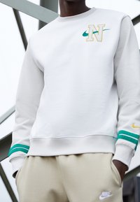 Vit sweatshirt med gul och teal Nike-logotyp samt gröna randiga detaljer på ärmarna, i kombination med ljusbeige byxor.