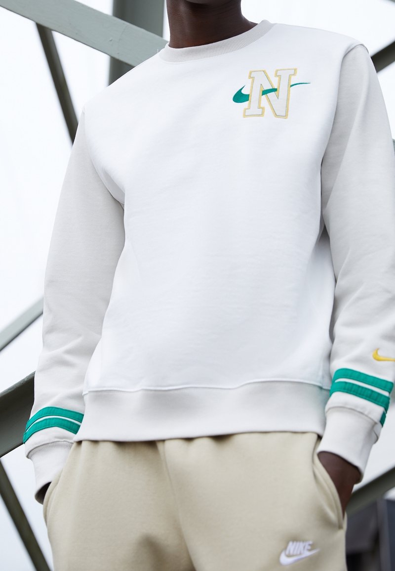 Vit sweatshirt med gul och teal Nike-logotyp samt gröna randiga detaljer på ärmarna, i kombination med ljusbeige byxor.