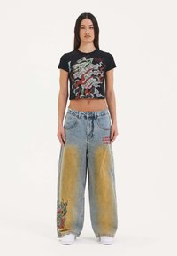 Black cropped t-shirt with graphic design, paired with oversized light blue denim jeans featuring yellow fades and embroidered dragon detail.