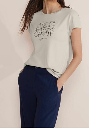 Kobieta ubrana w jasnobeżowy t-shirt z napisem "Adore Nature Create" i granatowe spodnie, stojąca z rękami w kieszeniach na jednolitym tle.