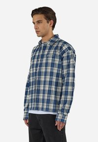 Jeune homme portant une chemise à carreaux bleue et blanche sur un t-shirt blanc et un pantalon noir, debout devant un fond uni.