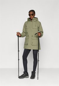 Olivgrüne Parka mit Reißverschluss und Knöpfen, zwei Brusttaschen, verstellbarer Kapuze und Kordelzug in der Taille. Schwarze Leggings und Wanderschuhe.