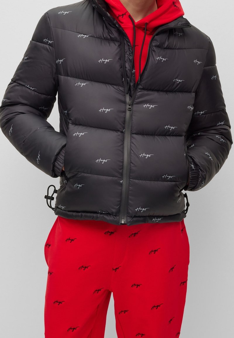 Czarna pikowana kurtka z zamkiem z przodu, elastycznymi mankietami i powtarzającym się nadrukiem logo. Noszona na czerwonym hoodie i dopasowanych spodniach.