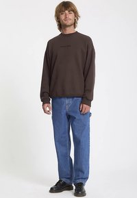 Felpa marrone con scollo rotondo, con piccolo logo. Abbinata a jeans denim blu e scarpe nere. Texture liscia, vestibilità comoda.