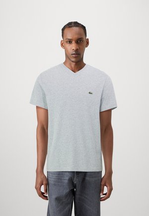 Camiseta gris de cuello en V hecha de tela suave con mangas cortas, que presenta un pequeño logo verde de Lacoste en el lado izquierdo del pecho. Lleva puesta unos jeans grises.