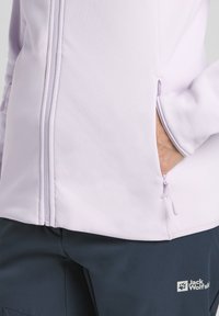 Veste zippée lilas clair avec poches latérales et une finition texturée, associée à un pantalon bleu marine foncé arborant un logo blanc.