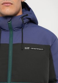 Plavi puffer jakna s crnom horizontalnom prugom, zelenim patentnim zatvaračem, podesivim kapuljačom i EA7 Emporio Armani logom na prsima.
